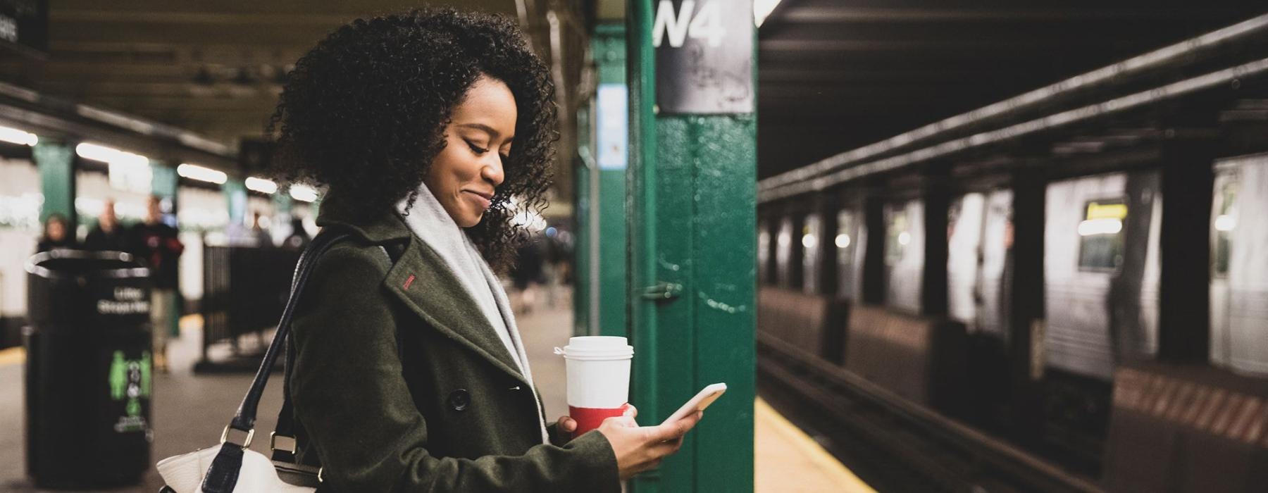 woman with a cup of coffee texts on subway station platform
